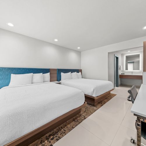 A modern hotel suite shows two beds with blue headboards, a vanity and kitchenette area, and recessed ceiling lights.