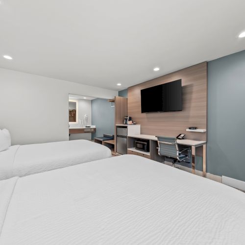 A modern hotel room with white beds, a desk setup, and a wall-mounted flat-screen TV under recessed lights.
