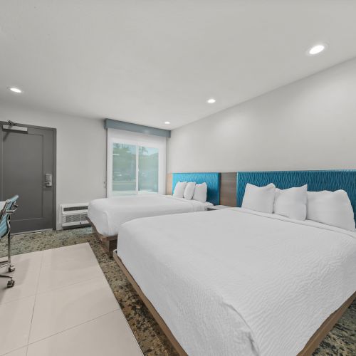 Modern hotel room with two white beds, blue headboards, a kitchenette counter, and a dark door under bright ceiling lights.