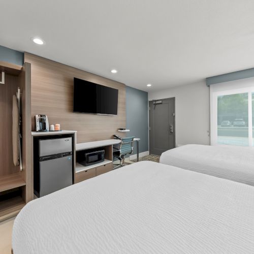 A modern hotel room with twin beds, a flat-screen TV, mini-fridge and coffee station, and a window with blinds.