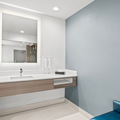 A modern bathroom vanity with sink and mirror, soap/tissue dispensers, a small towel, and a view into a shower area.