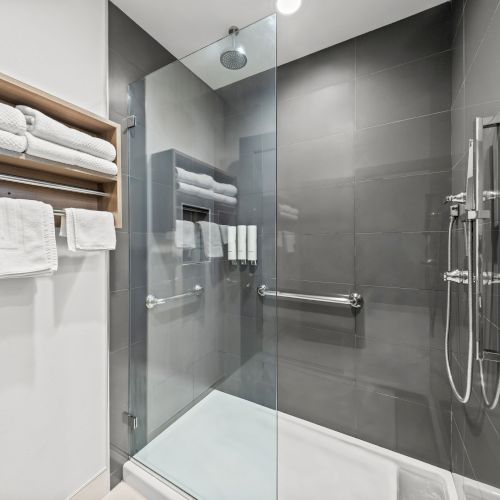 A modern bathroom shower with a glass enclosure, rainfall showerhead, chrome fixtures, and folded white towels on a wooden shelf.