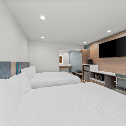 A bright hotel room with two beds, a desk and chair, a coffee maker, and a wall-mounted flat-screen TV.