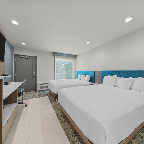 A modern hotel room with two beds, a flat-screen TV, desk cabinets, and a door and window with blinds.