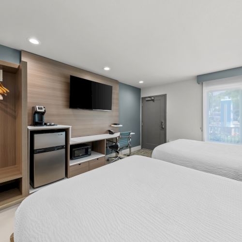 Modern hotel room with twin beds, a wall TV, desk area, and coffee station near a large window.