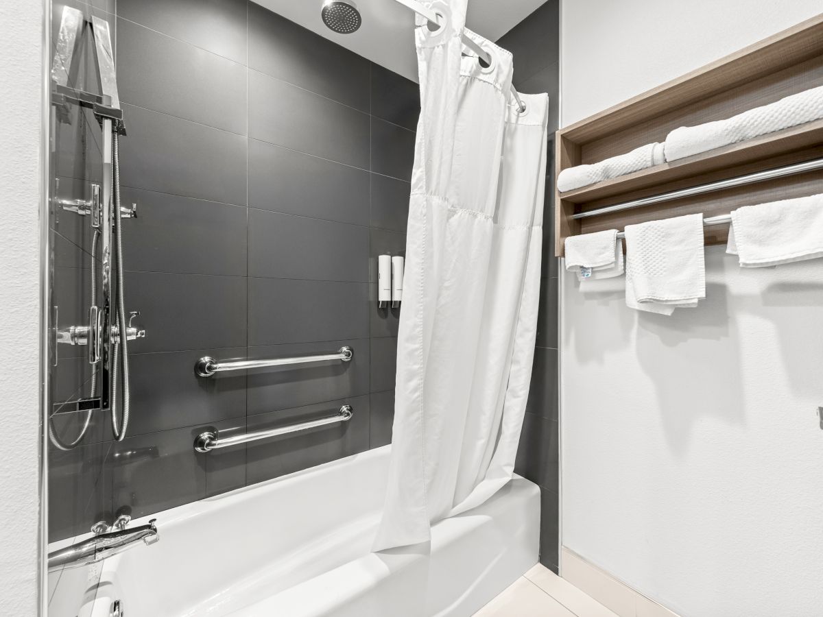 A modern bathroom with a bathtub, glass shower panel, white shower curtain, towels on a shelf, and a ceiling showerhead.